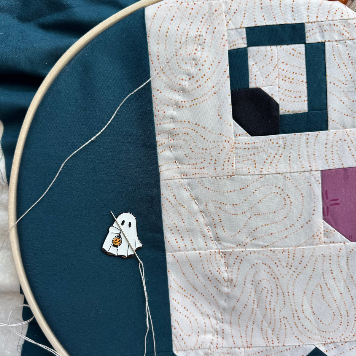 A close-up of a handmade quilt in progress, featuring a white and teal ghost pattern with orange dotted topographic lines, a dark teal fabric background, and a small embroidered white ghost with a pumpkin patch.