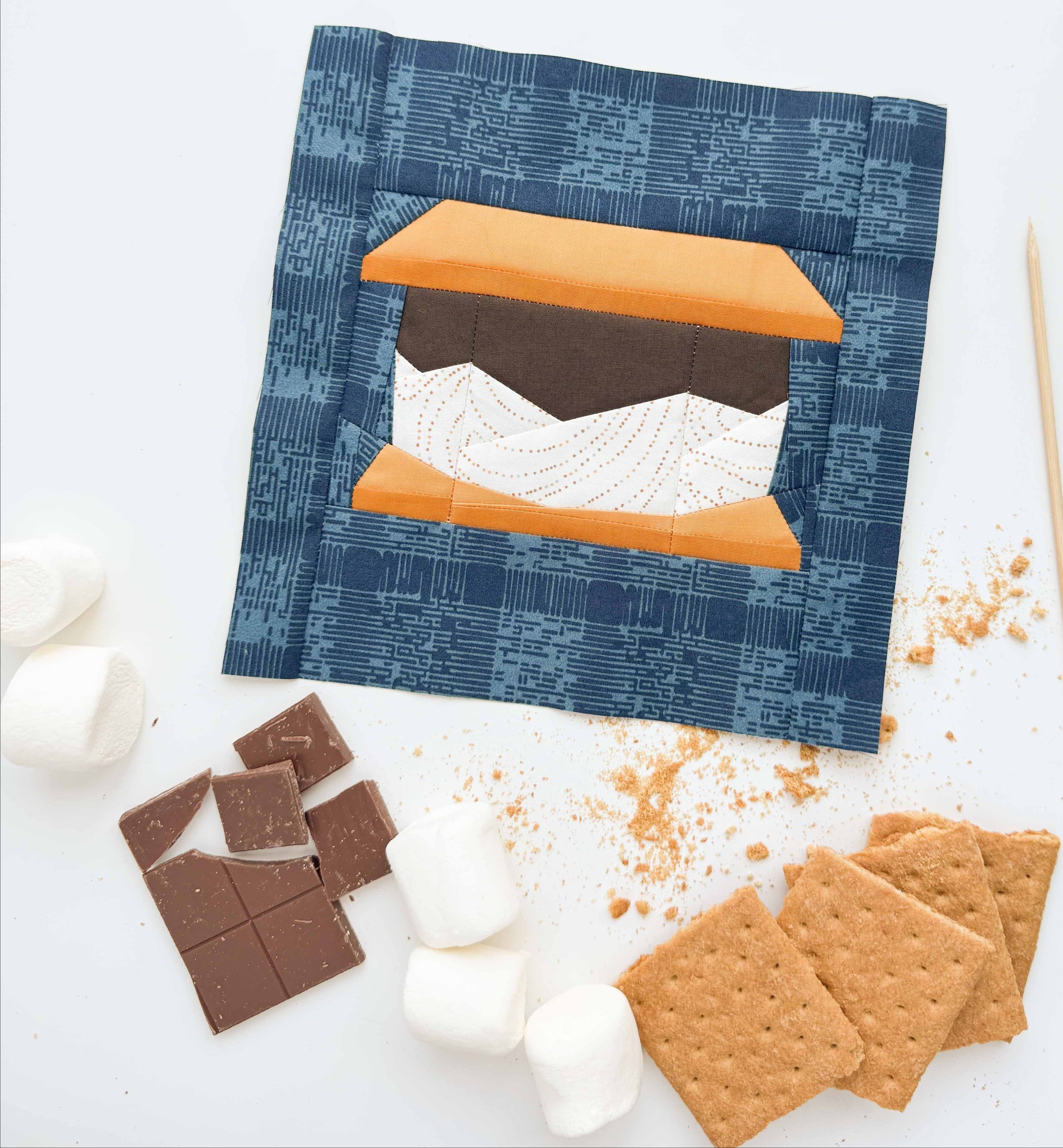 S'mores-themed quilt block with graham crackers, chocolate, marshmallows, and sticks on a white background.